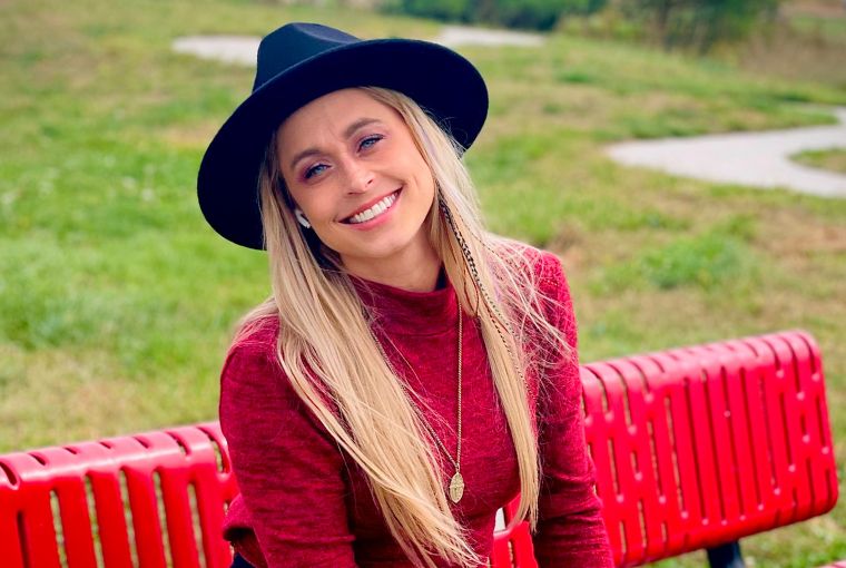 Successful salon owner and solo stylist Jenna Morehouse sits on a bright red bench in a park wearing a black hat and red top